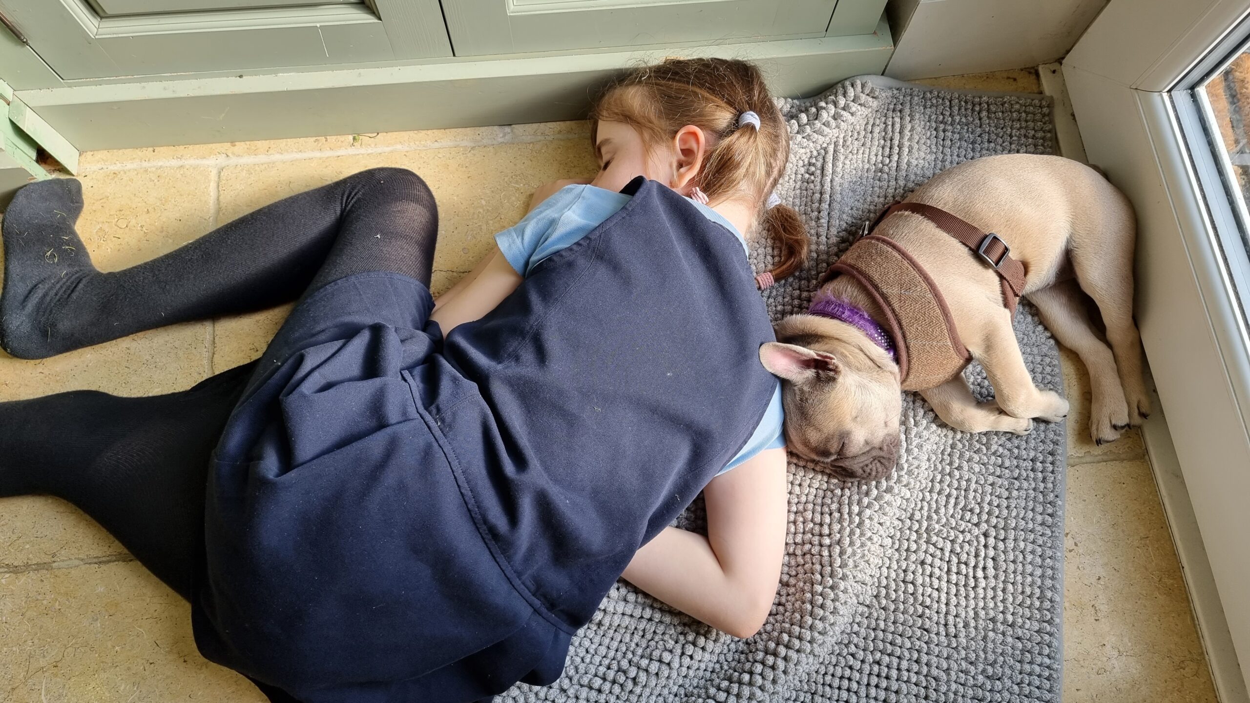 Full image of A young girl asleep on a stone floor, her head on a doormat, napping alongside a French Bulldog.