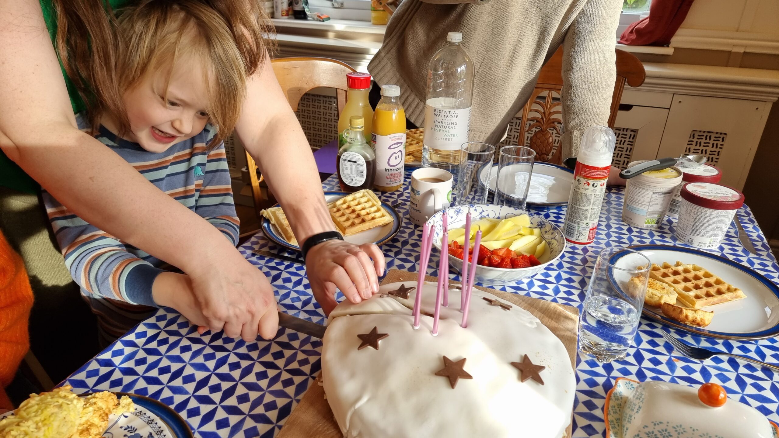 Full image of A boy in blue-and-brown striped pyjamas, on a table stacked with plates of waffles, slices into a birthday cake with six candles.
