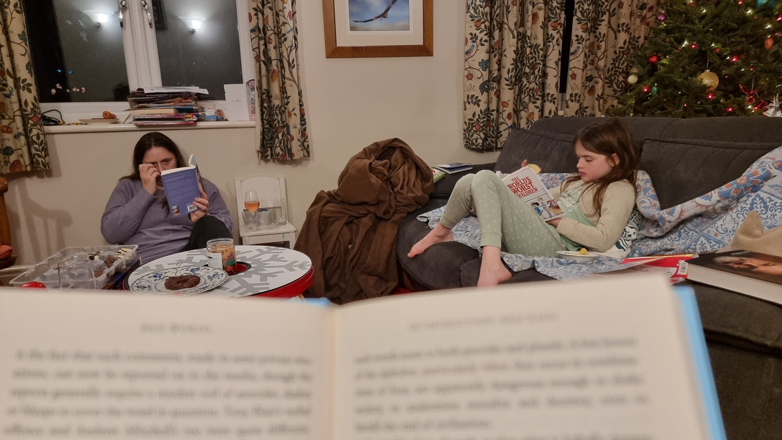 Full image of Family members read books in a living room, seen over the top of the pages of an (out of focus) book in the foreground.