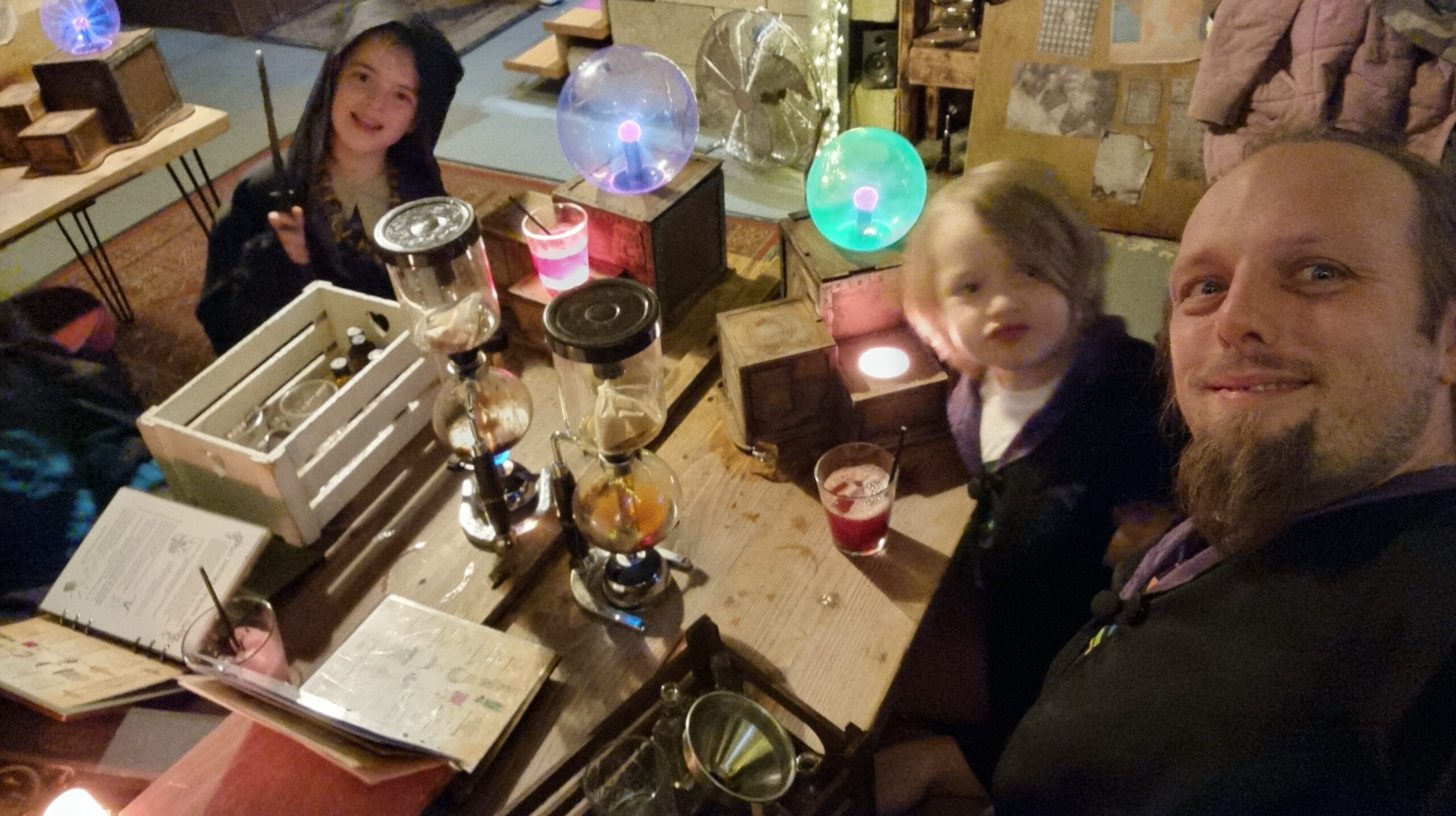 Full image of Dan, wearing wizard's robes alongside two similarly-dressed children, makes cocktails using a selection of bizarre-looking laboratory equipment.