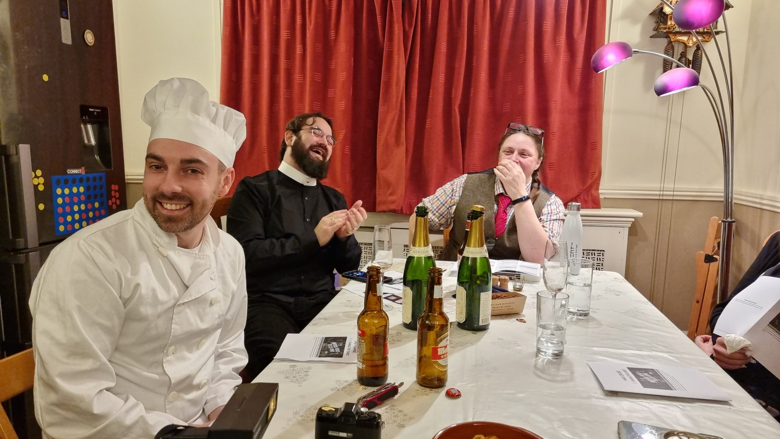 Full image of Guests dressed as a chef, a priest, and a librarian sit around a dining table at a murder mystery party.