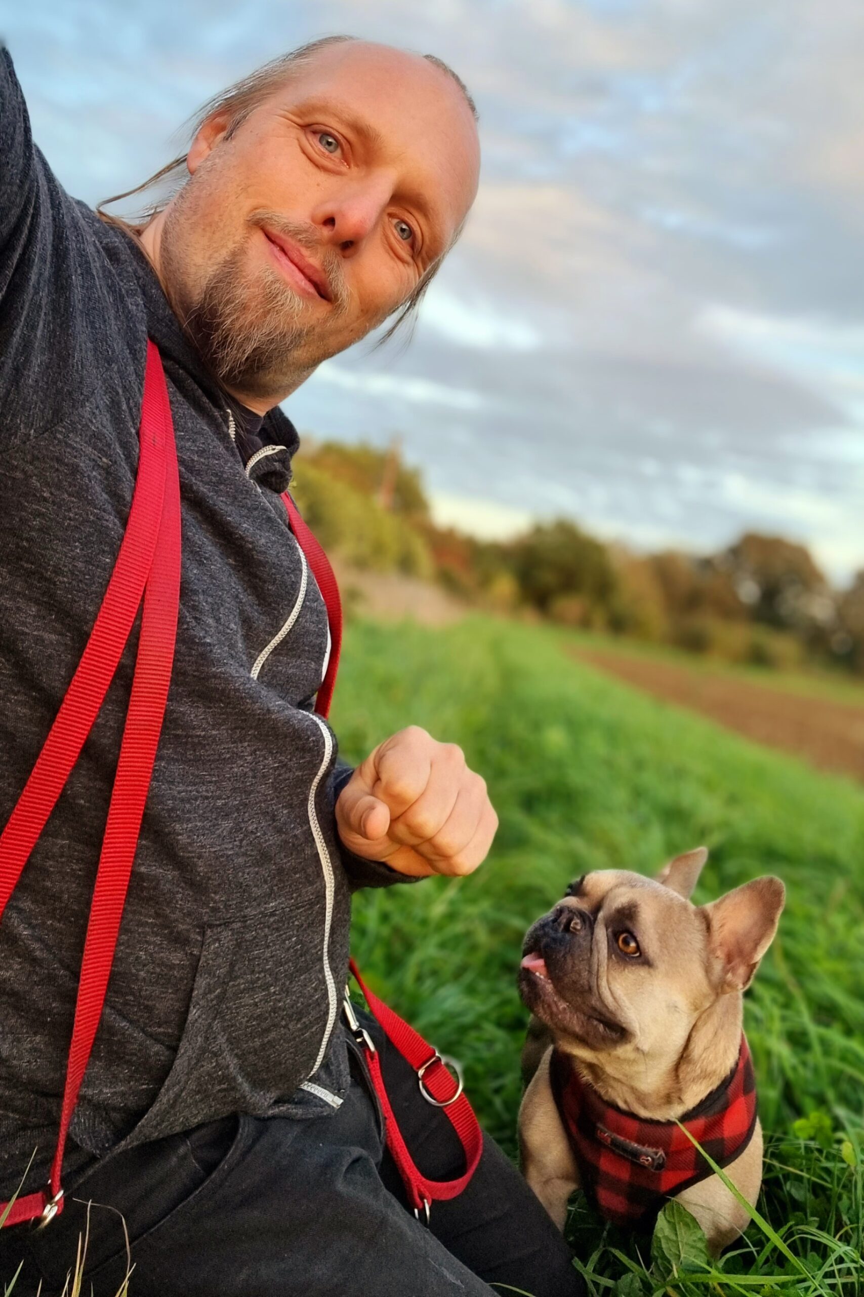 Full image of Dan kneels in the grassy verge of a recently-ploughed field in late-afternoon summer sunshine, a long red dog's lead hanging around his shoulders. By his side, a champagne French Bulldog looks with anticipation up at him (or possibly at his closed fist).