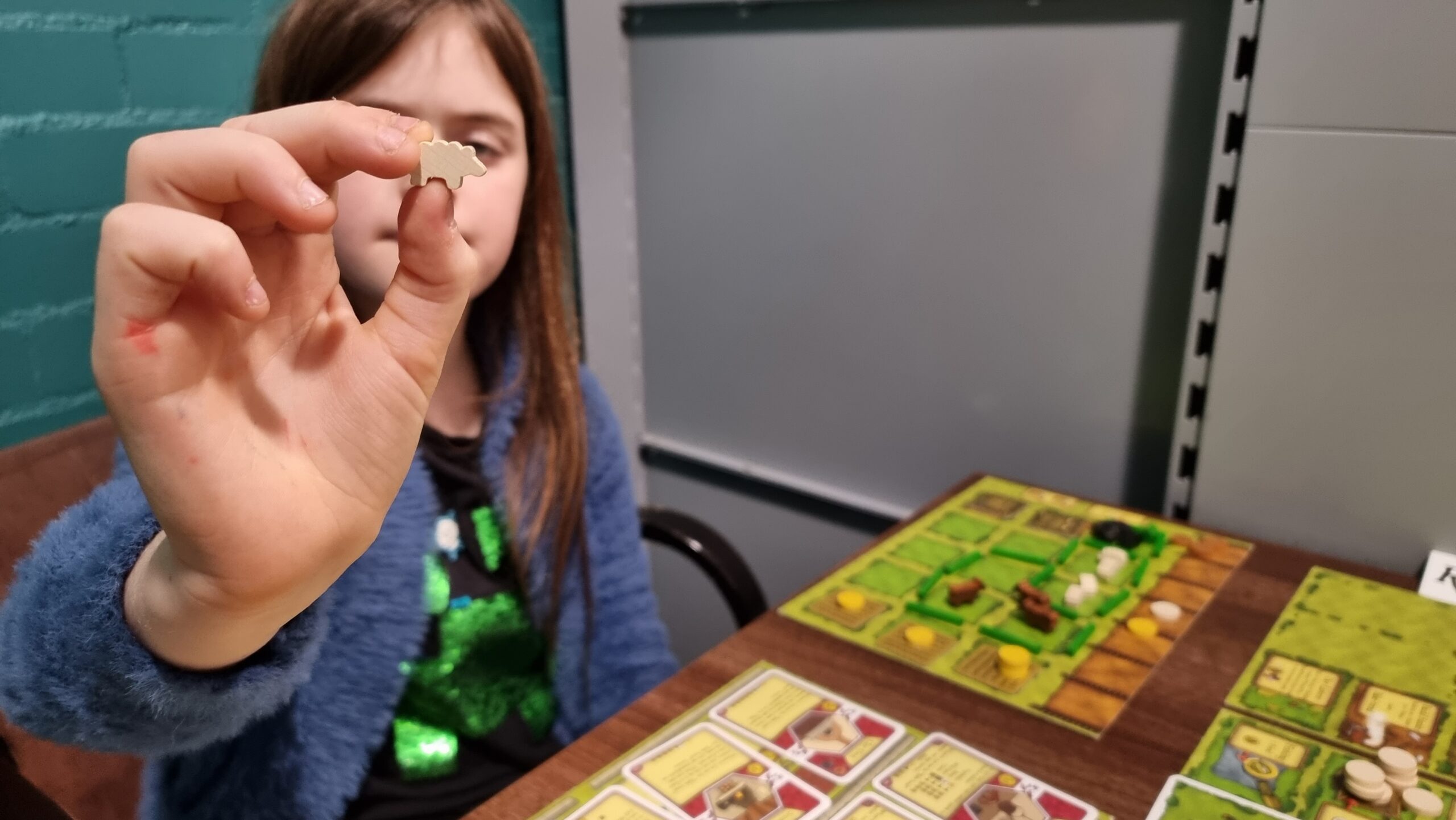 Full image of A girl, sat in front of an Agricola farmyard board, holds up a "sheeple" (small wooden sheep game piece) for the camera.