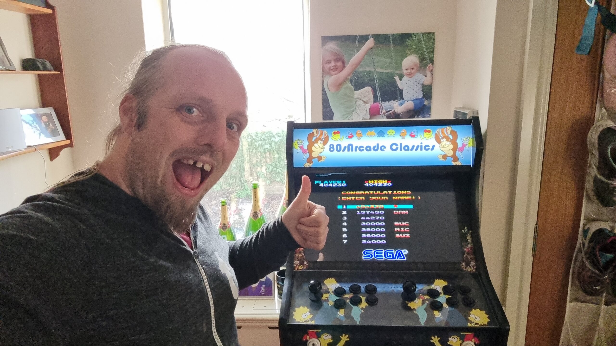 Full image of Dan with his thumbs-up in front of the high-score table (with the top-ranking spot about to be filled) of Wonder Boy, on a generic "80s Arcade Classics" arcade cabinet.