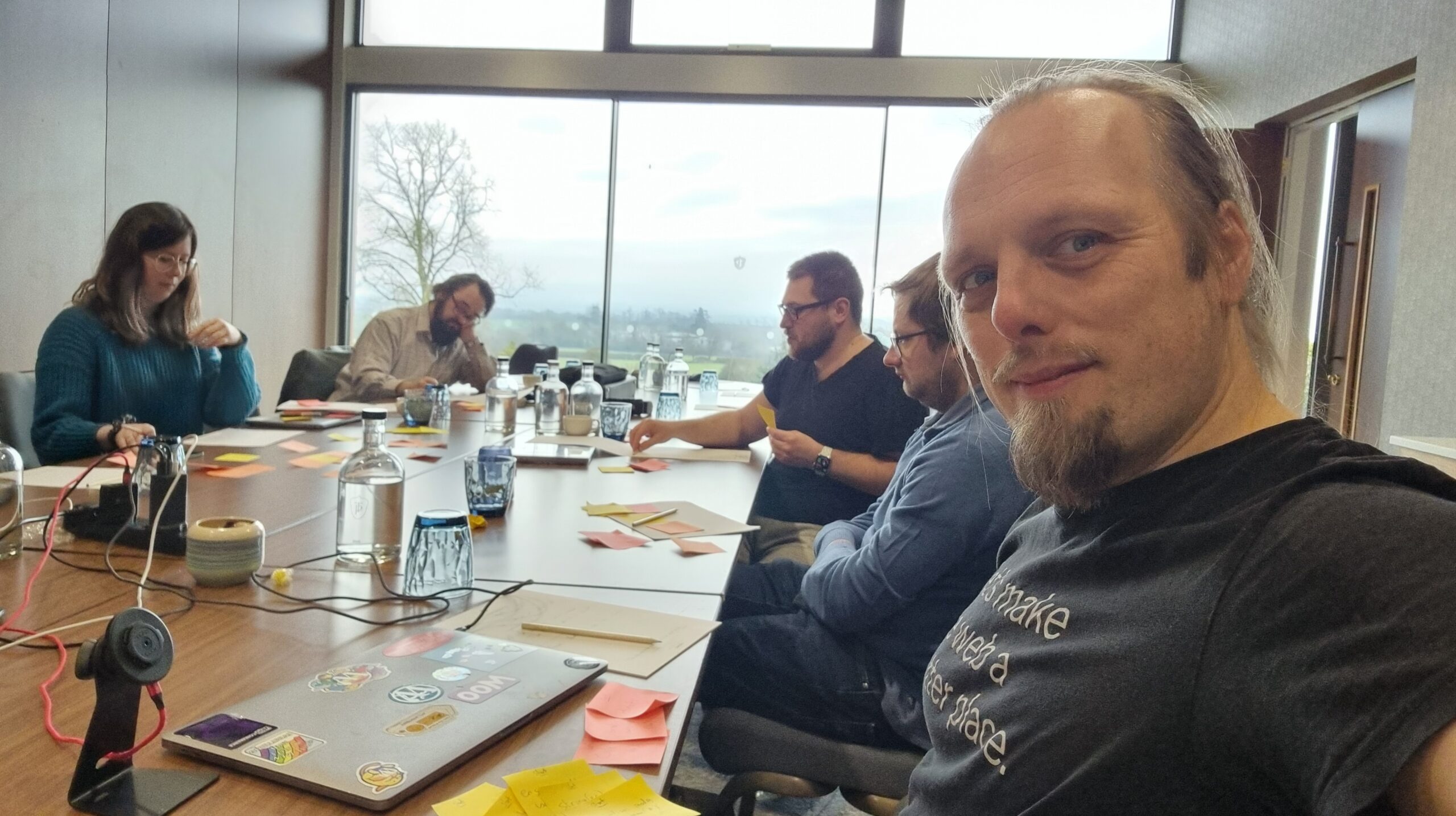 Full image of Dan sits at a boardroom table in an airy, bright room. He's wearing an Automattic t-shirt that reads "Let's make the Web a better place." In the background, several other people discuss a pile of post-it notes that have begun to pile up on the table.