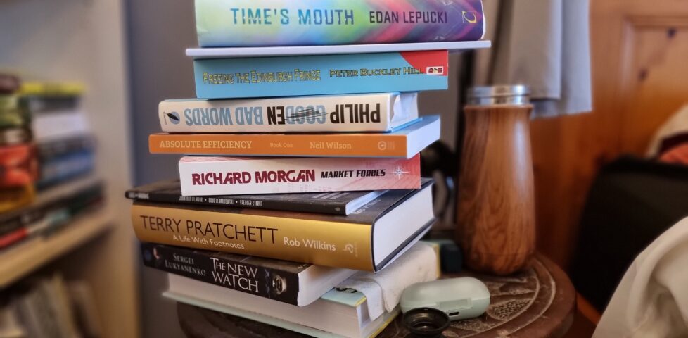 A pile of books on a round-topped wooden bedside table. From top to bottom: Time's Mouth by Edan Lepucki, a slim book whose spine doesn't show a name, Bad Words by Philip Gooden, Absolute Efficiency: Book One by Neil Wilson, Market Forces by Richard Morgan, Kids on Brooms by Jonathan Gilmour, Doug Levandowski, and Spenser Starke, Terry Pratchett: A Life With Footnotes by Rob Wilkins, The New Watch by Sergei Lukyanenko, and a book whose spine is turned away from the camera.