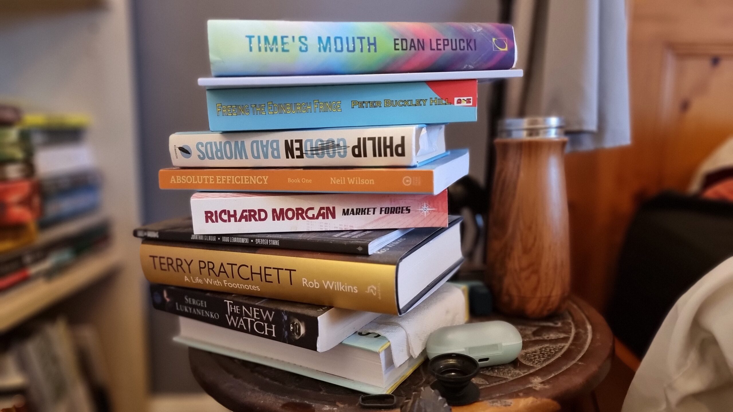 Full image of A pile of books on a round-topped wooden bedside table. From top to bottom: Time's Mouth by Edan Lepucki, a slim book whose spine doesn't show a name, Bad Words by Philip Gooden, Absolute Efficiency: Book One by Neil Wilson, Market Forces by Richard Morgan, Kids on Brooms by Jonathan Gilmour, Doug Levandowski, and Spenser Starke, Terry Pratchett: A Life With Footnotes by Rob Wilkins, The New Watch by Sergei Lukyanenko, and a book whose spine is turned away from the camera.
