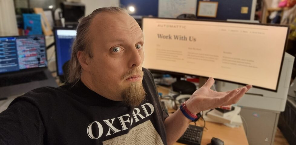 Dan, wearing an Oxford-branded t-shirt, shrugs and looks confused in front of a screen showing Automattic's "Work With Us" page.