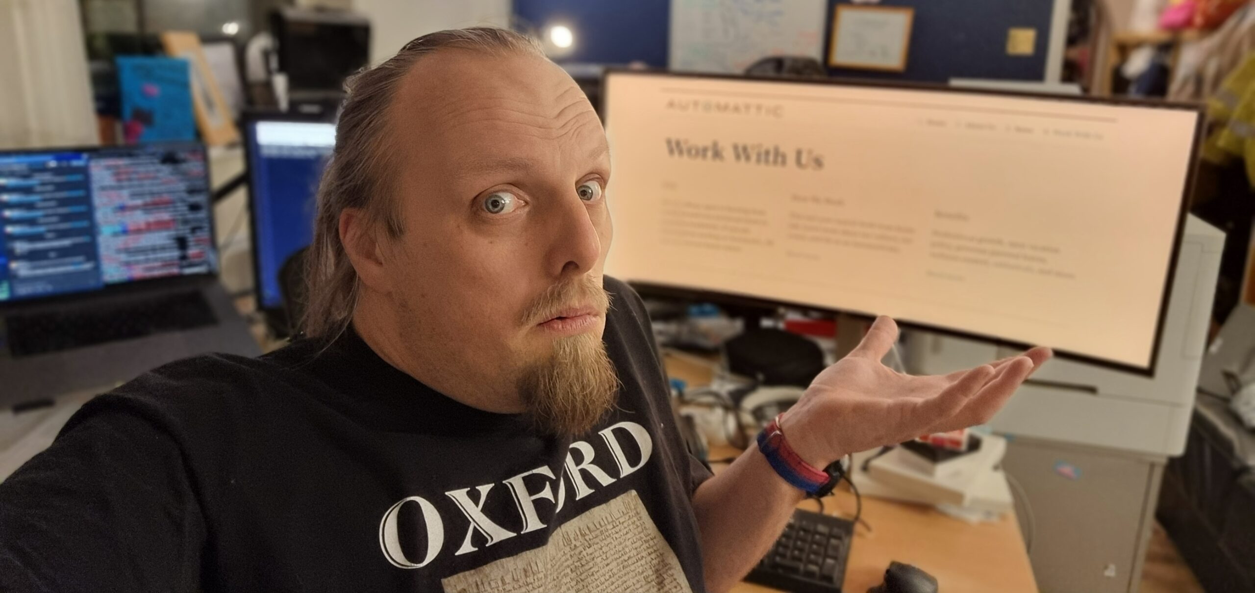 Full image of Dan, wearing an Oxford-branded t-shirt, shrugs and looks confused in front of a screen showing Automattic's "Work With Us" page.