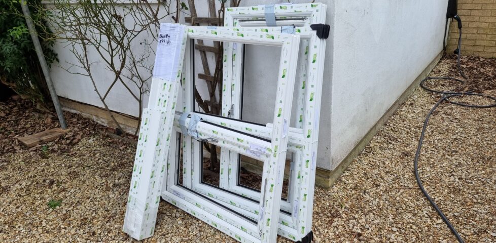 A pile of window frames, factory-fresh and covered with tape. propped against the side of a white house, on a gravel driveway.