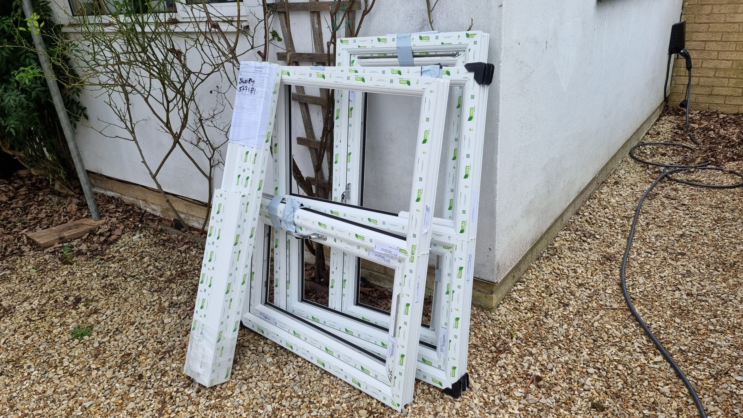 Full image of A pile of window frames, factory-fresh and covered with tape. propped against the side of a white house, on a gravel driveway.