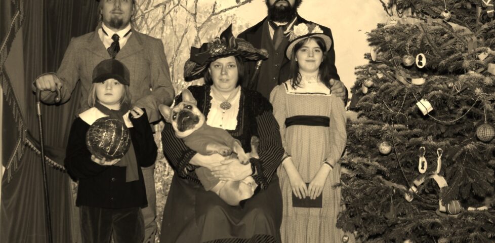 Posed sepia photograph of Dan, JTA, Ruth, and their children and dog, dressed in Victorian-era clothing, by a Christmas tree.