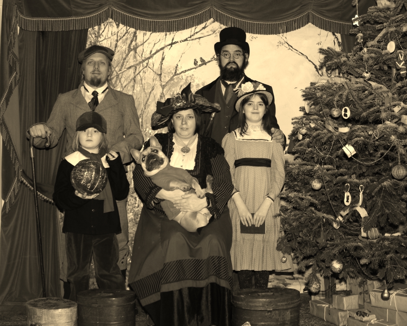 Full image of Posed sepia photograph of Dan, JTA, Ruth, and their children and dog, dressed in Victorian-era clothing, by a Christmas tree.