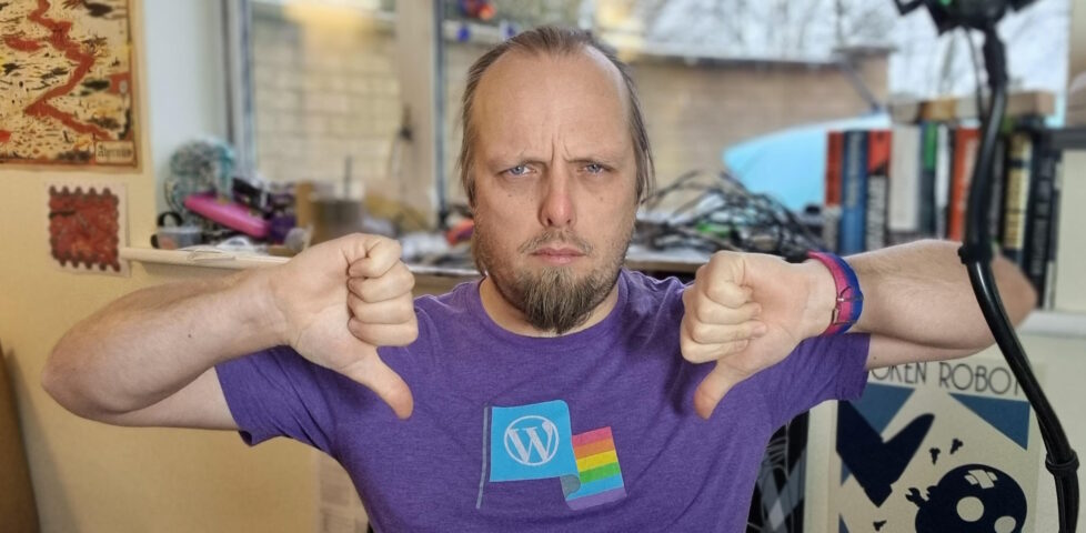 Dan, wearing a purple t-shirt with a WordPress logo and a Pride flag, sits in his home office and gives two "thumbs down" signs while frowning at the camera.