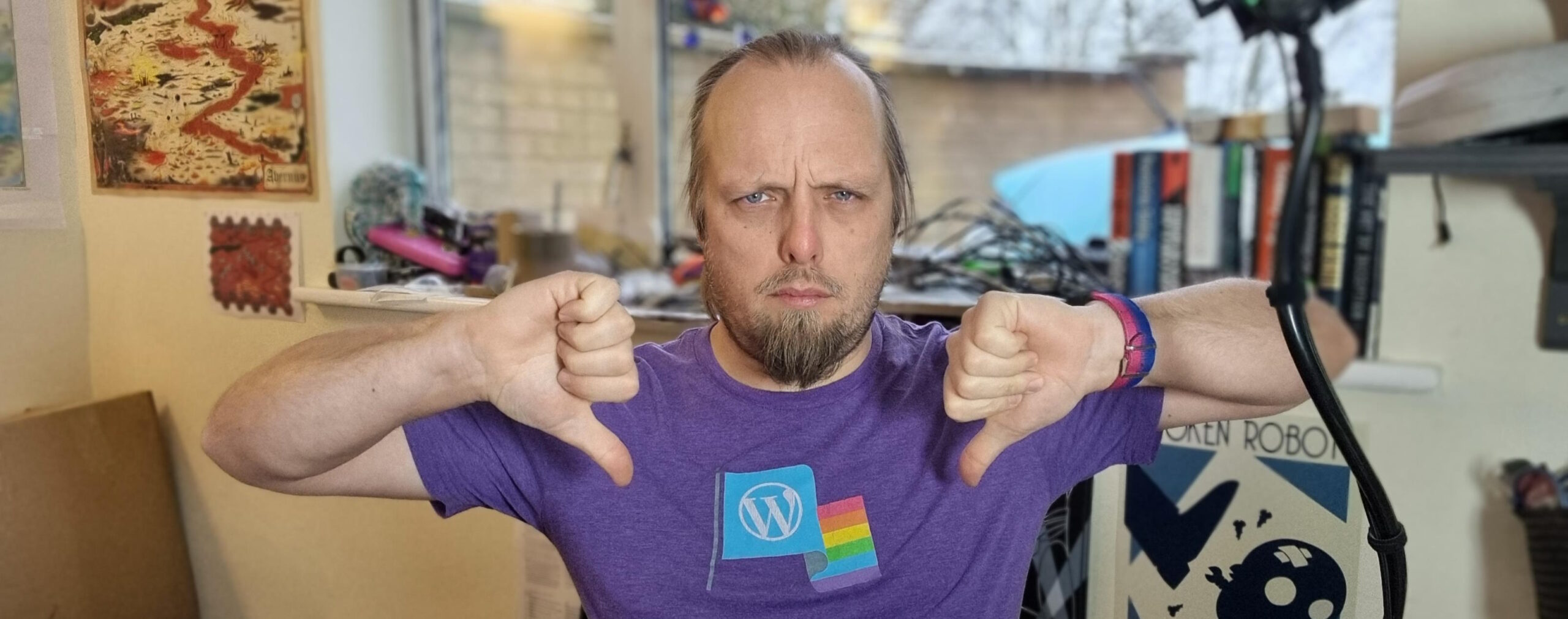 Full image of Dan, wearing a purple t-shirt with a WordPress logo and a Pride flag, sits in his home office and gives two "thumbs down" signs while frowning at the camera.