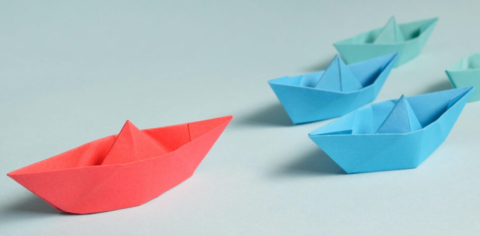 A flotilla of paper boats on a table: a red boat leads the way ahead of two blue and two green boats.
