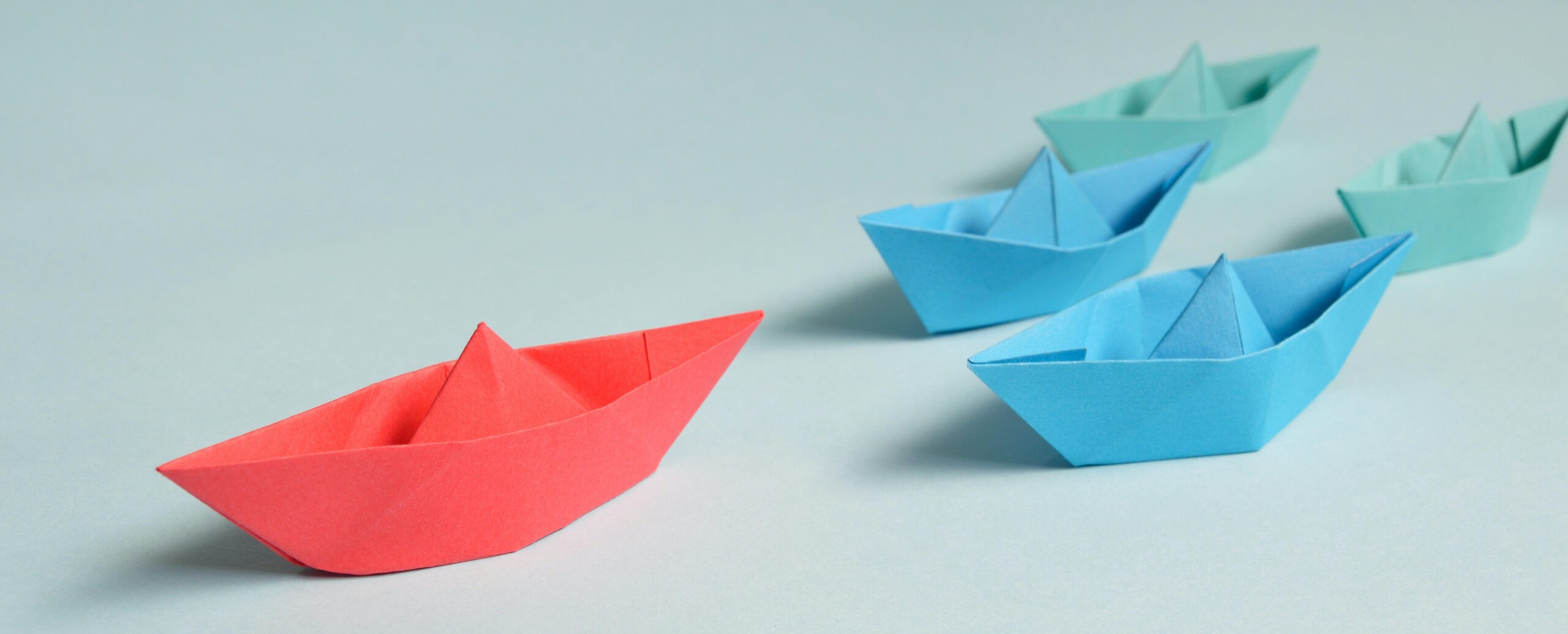 Full image of A flotilla of paper boats on a table: a red boat leads the way ahead of two blue and two green boats.