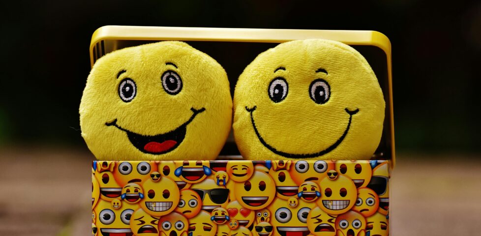 Two soft-toy emoji-style smiley faces peeping out of a small box which is decorated with smiley emojis.