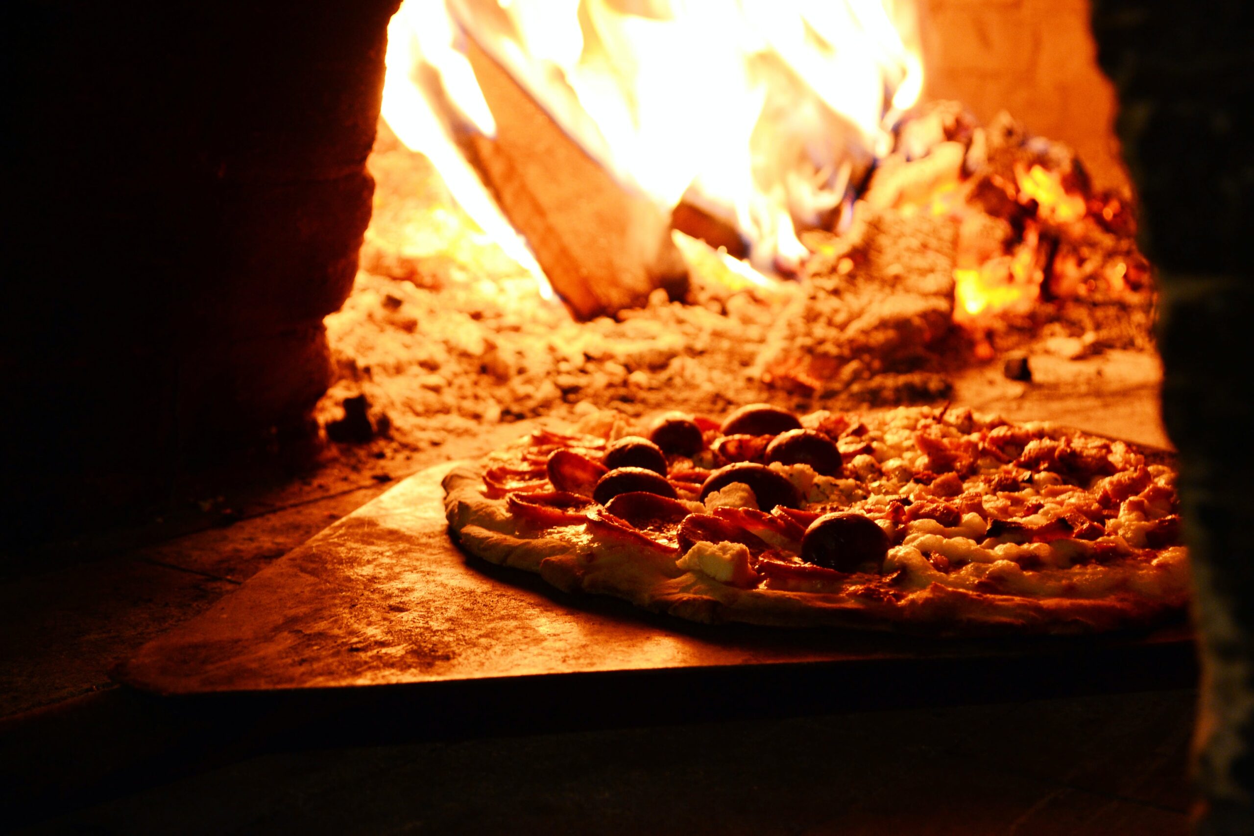 Full image of A pizza slides off a peel into a wood-fired oven. One half seems to have salami and olives, the other half perhaps just cheese and tomato.