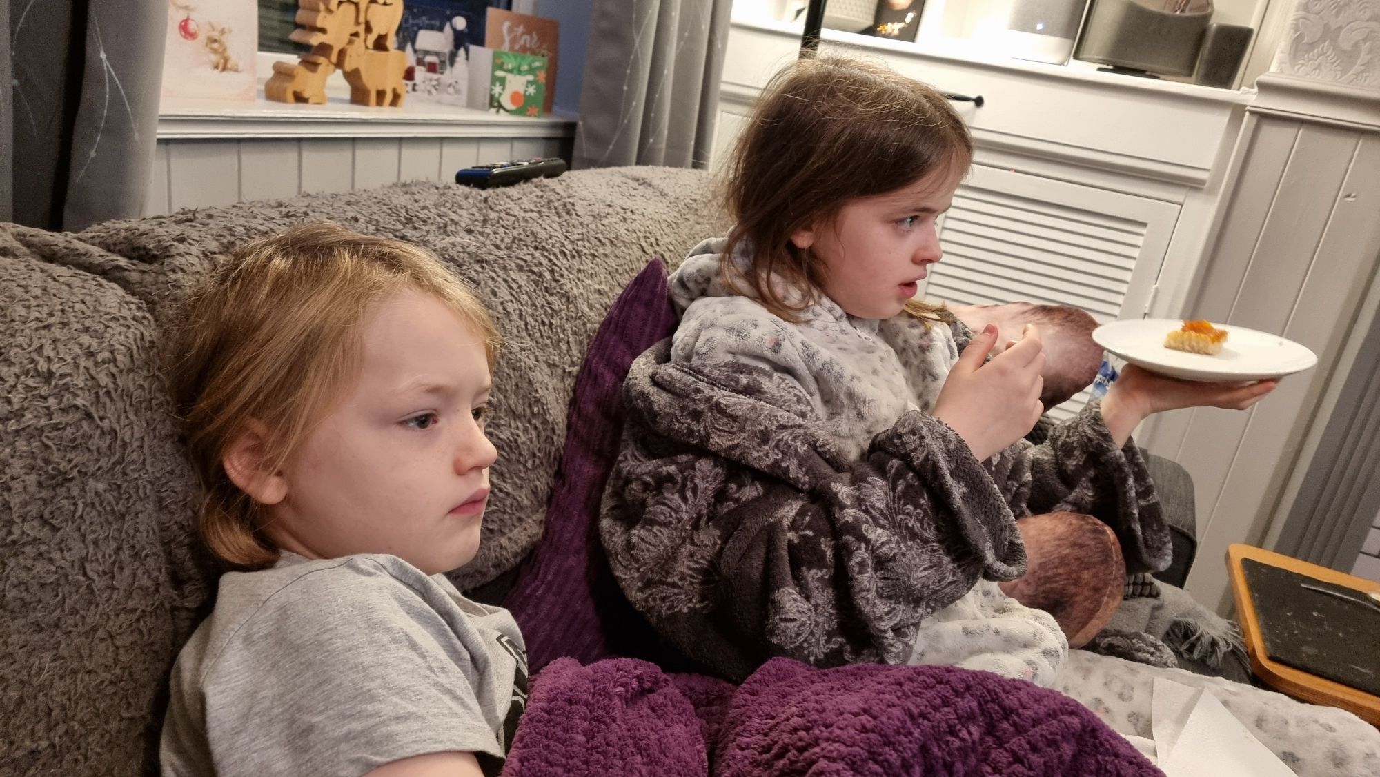 Full image of A young boy and a less-young girl sit on a sofa in pyjamas and dressing gowns.