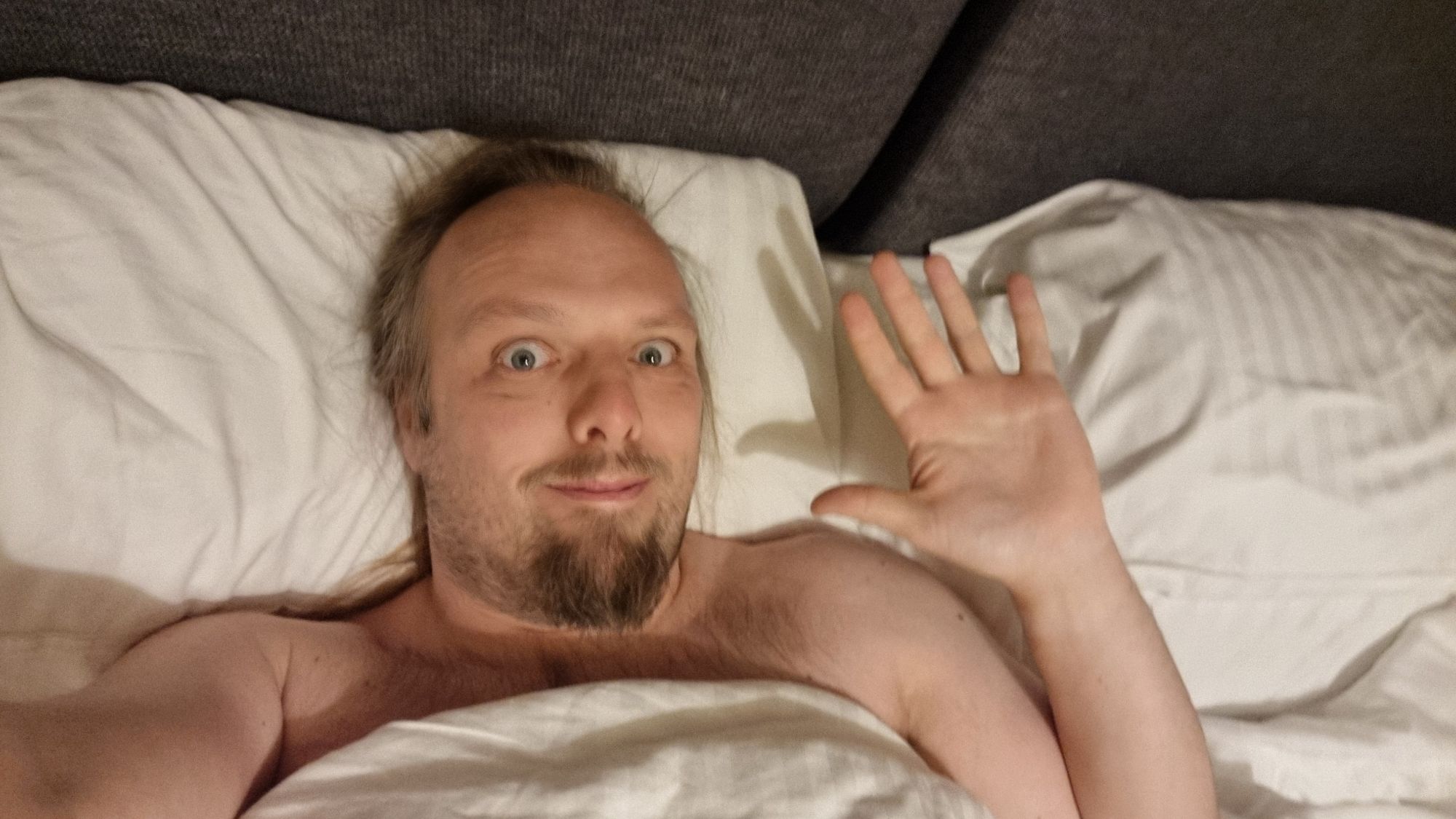 Full image of Dan waves, his head and shoulders peeping out from underneath a white duvet.