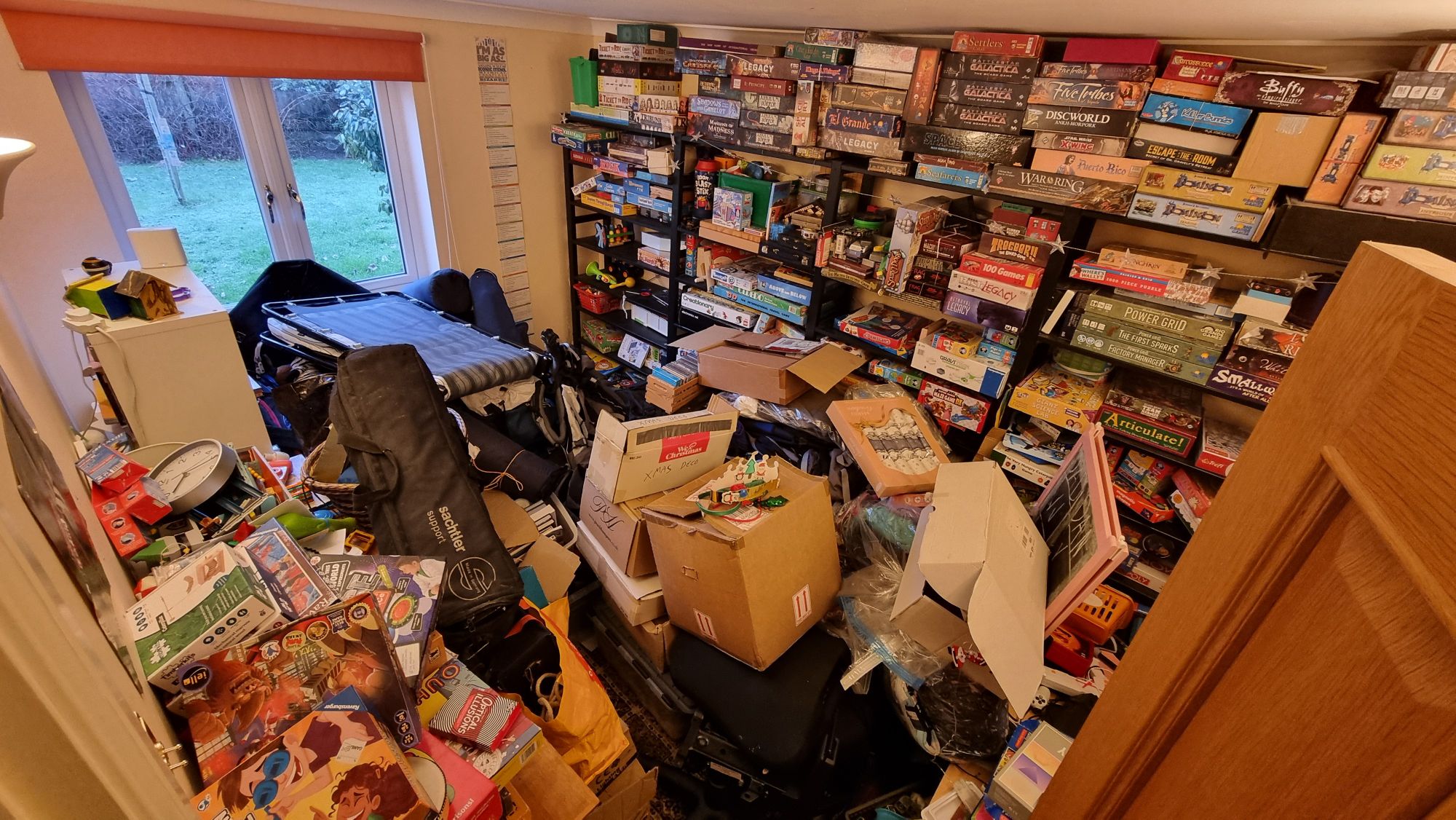 Full image of An extremely cluttered room: shelved board games line the walls, heaps of boxes fill all the floor space and are stacked chest-high.