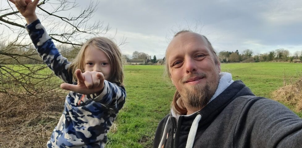 Dan, wearing a grey hoodie, stands in a meadow alongside a boy who's throwing "devil horns" hand signs.