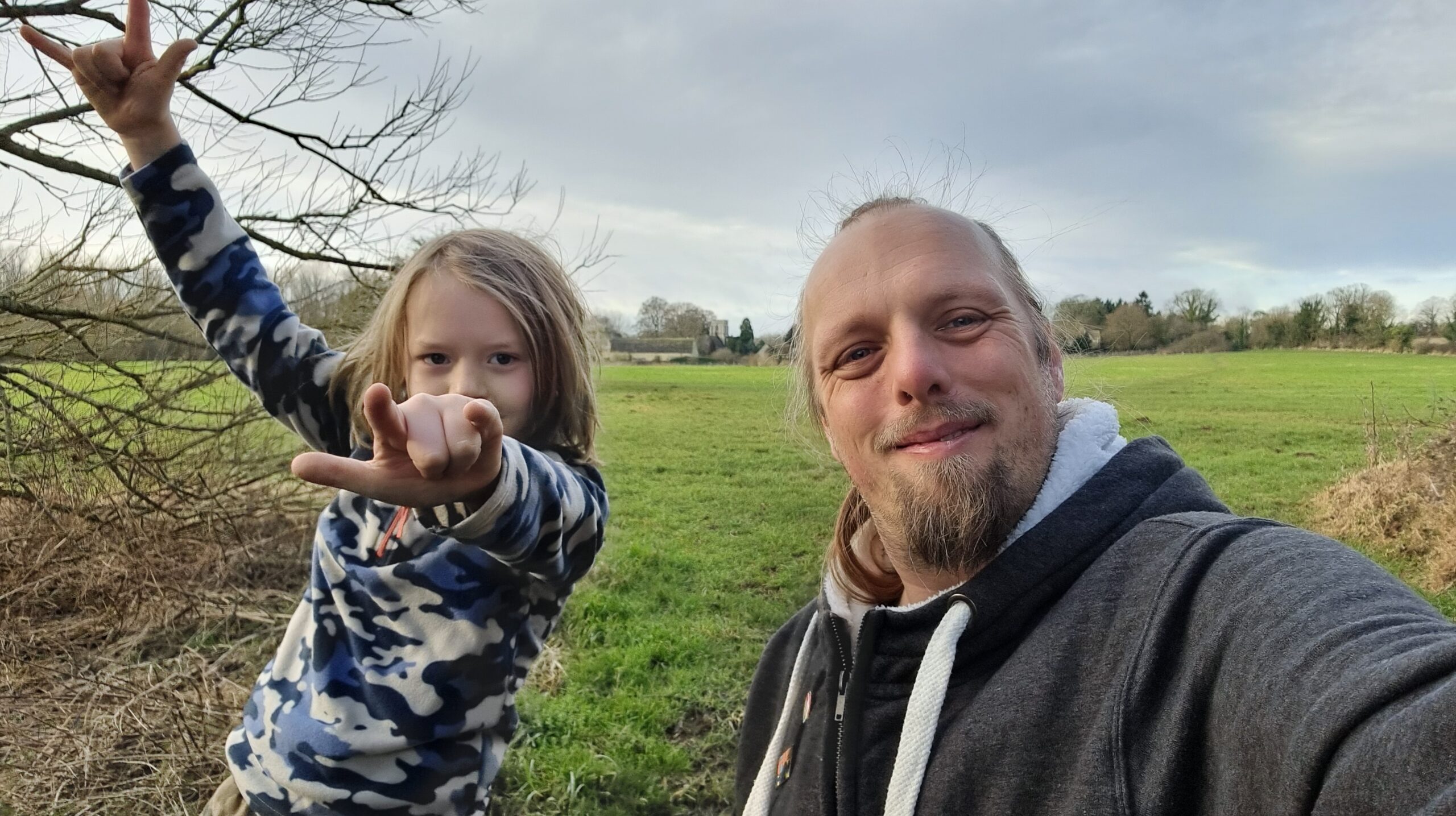 Full image of Dan, wearing a grey hoodie, stands in a meadow alongside a boy who's throwing "devil horns" hand signs.