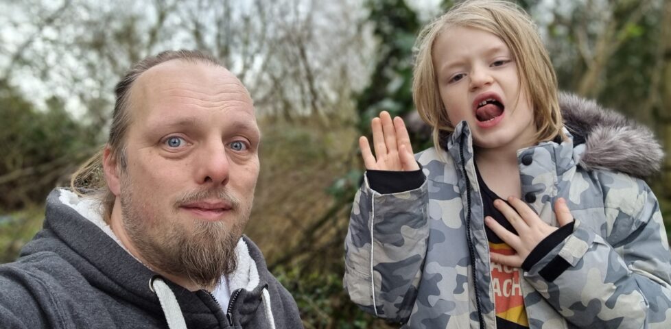 Dan, in a grey jumper, stand beside a 7-year-old boy wearing a camo jacket, in a lightly wooded area.