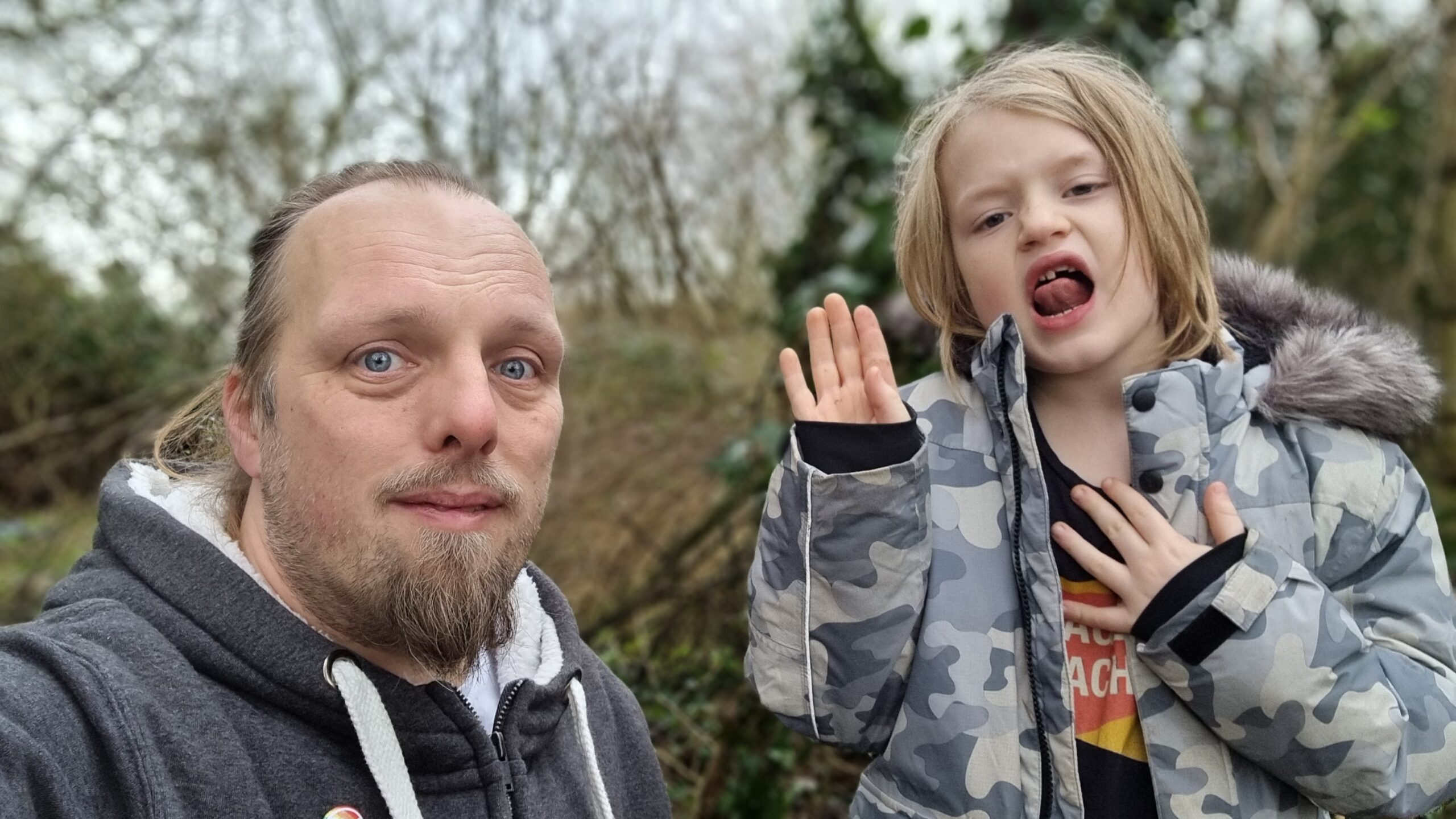 Full image of Dan, in a grey jumper, stand beside a 7-year-old boy wearing a camo jacket, in a lightly wooded area.