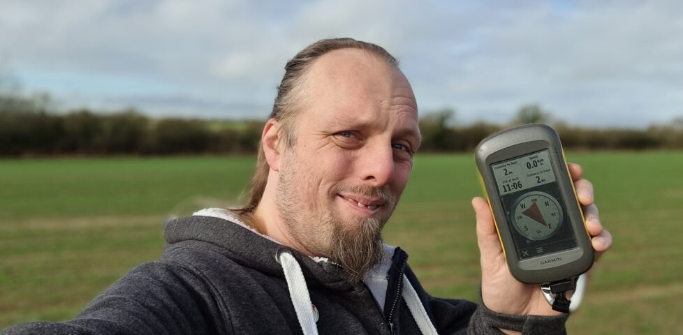 Dan, smiling, holding up his GPSr, in a field.