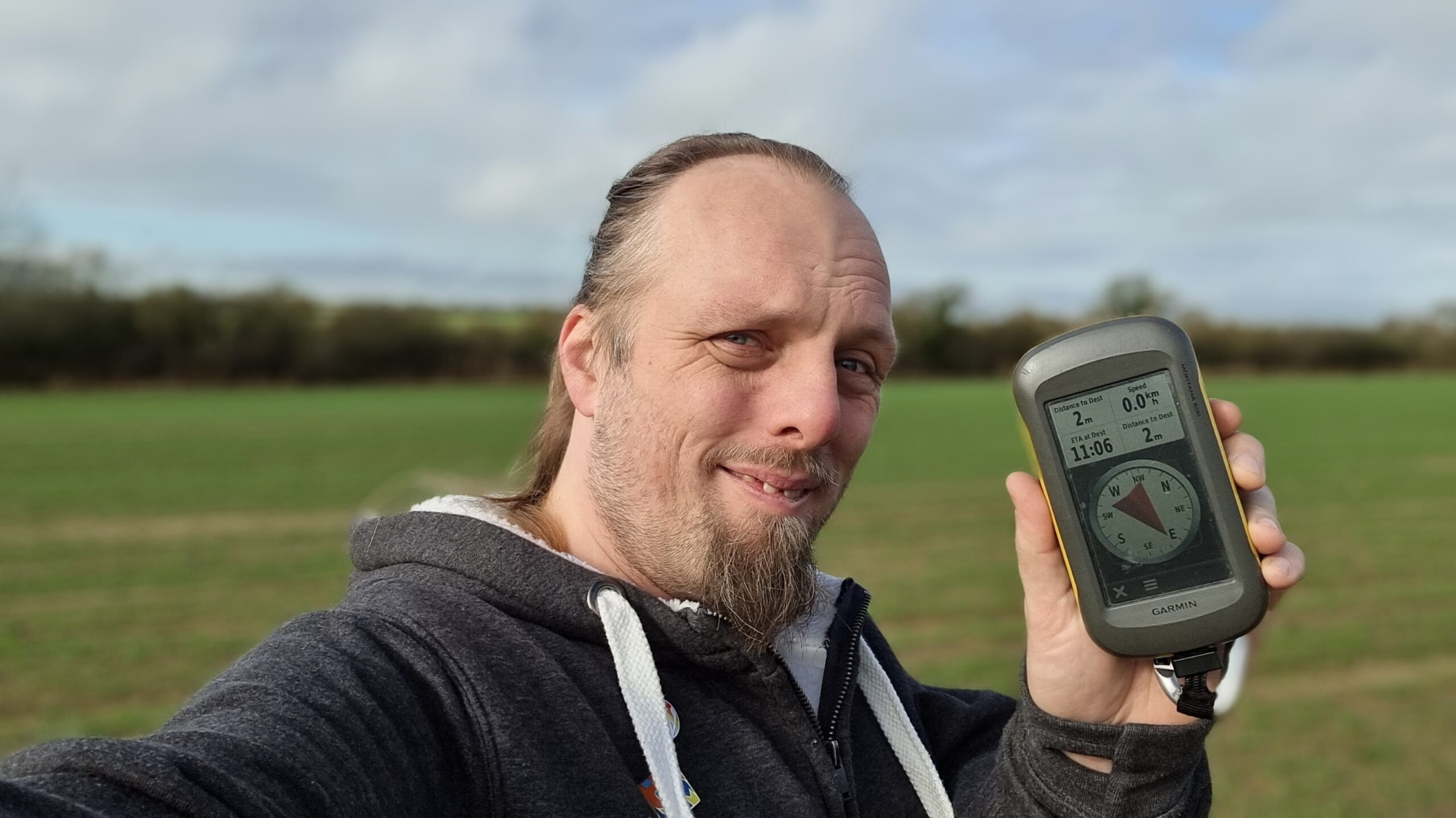 Full image of Dan, smiling, holding up his GPSr, in a field.