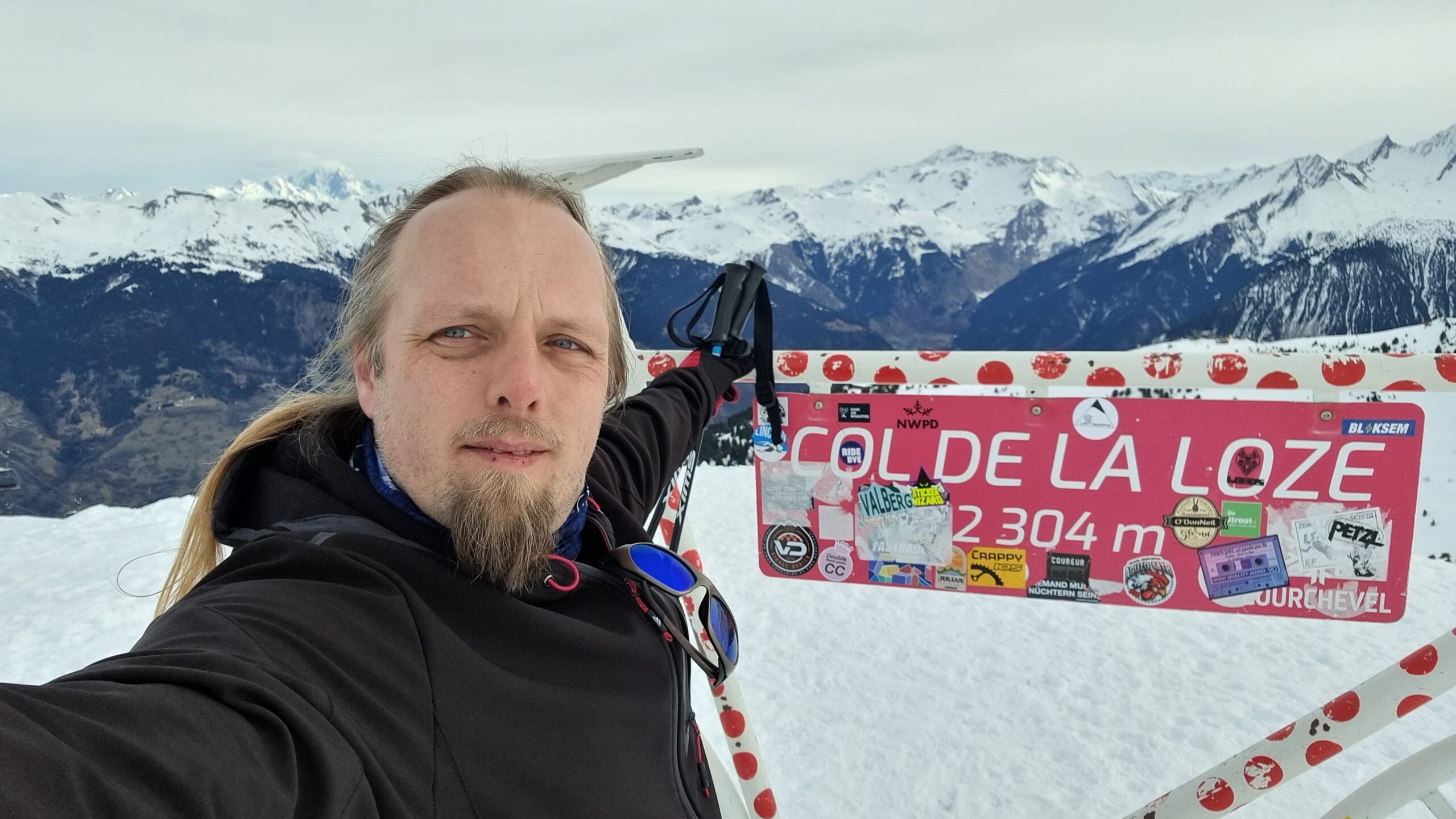 Full image of Dan, in a wintery mountainous landscape, poses for a selfie by a sculpture of a bicycle marking part of the route of the Tour de France. 