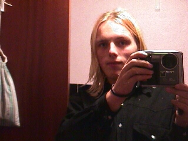 A young white man with platinum blonde hair holds up a large, boxy digital camera.