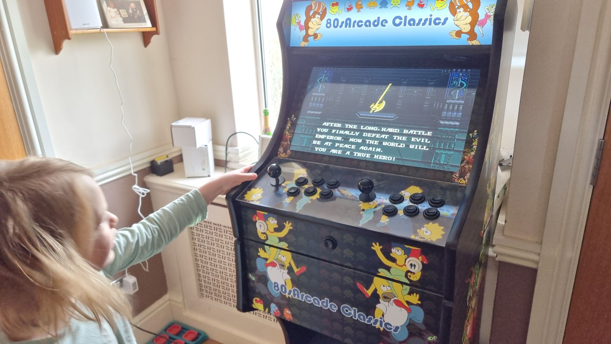 Full image of A young boy in green pyjamas stands alongside a video games cabinet. The screen shows 16-bit graphics of a golden axe, below which the text reads: "After a long, hard battle you finally defeat the evil emperor. Now the world will be at peace again. You are a true hero!"