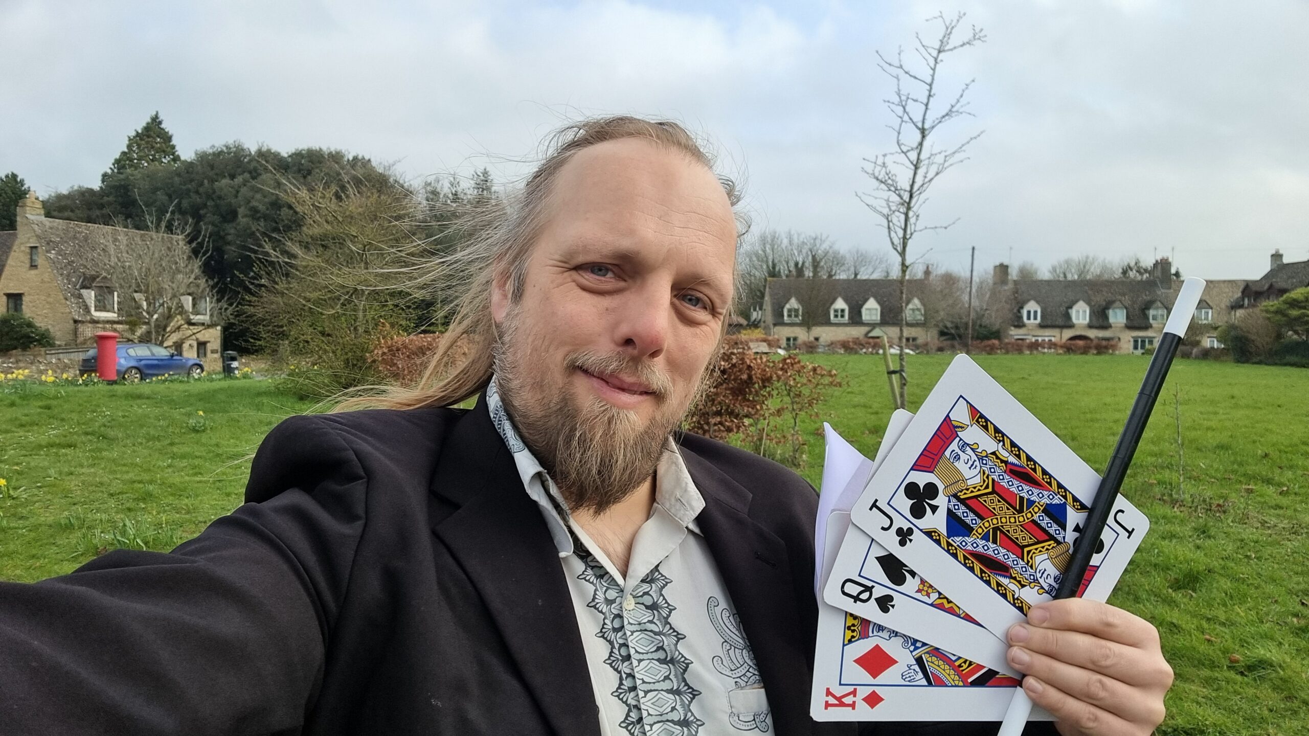 Dan, a white man with long hair (tied back) and a beard, stands in a Cotswolds-esque village green wearing a black jacket and holding three large novelty playing cards and a magic wand.