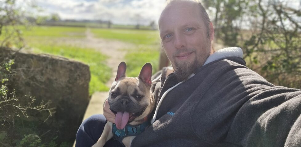 Dan sits on a footbridge in a field, a French bulldog on his lap, in spring morning sunshine.