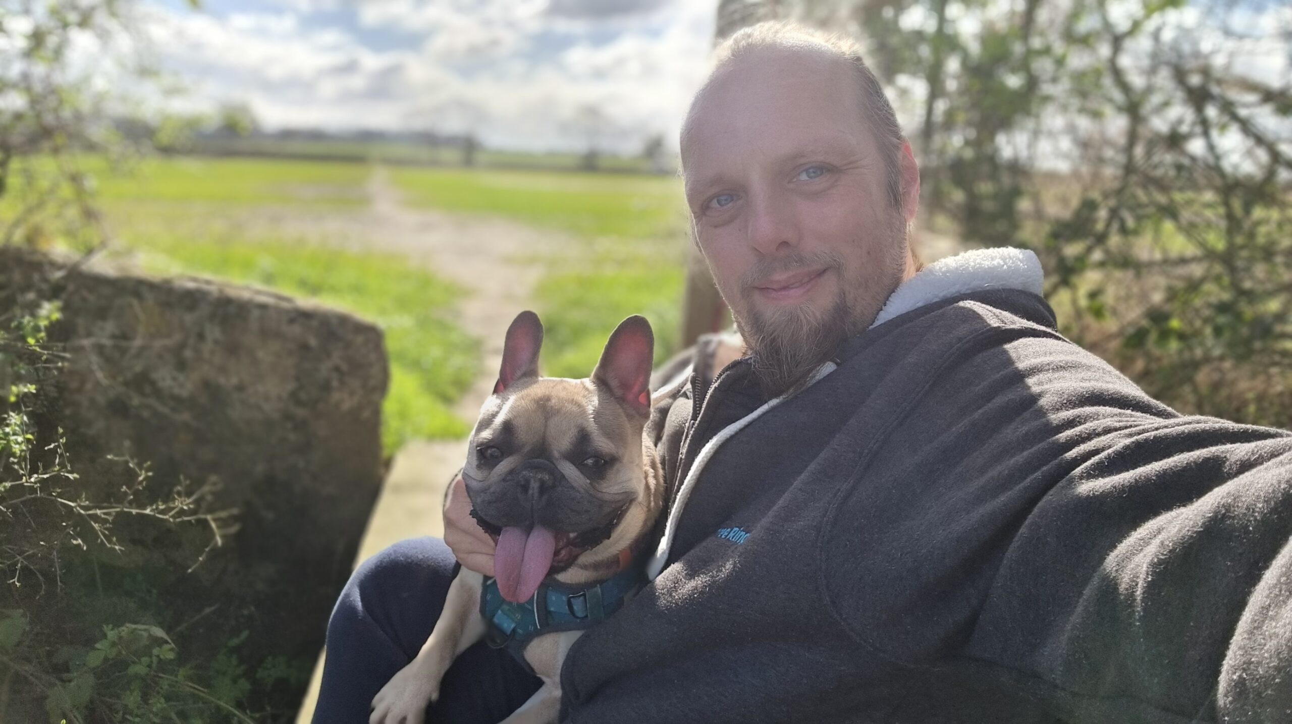 Full image of Dan sits on a footbridge in a field, a French bulldog on his lap, in spring morning sunshine.