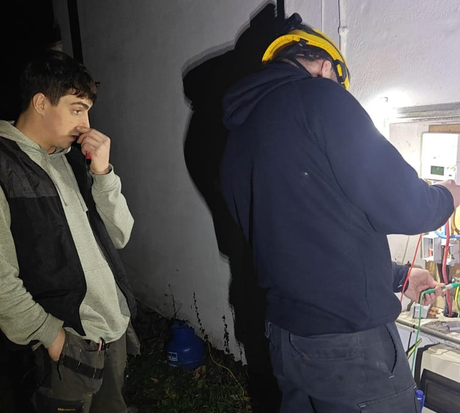 Two electricians, one in a hard hat, look in an outdoor metering cupboard by torchlight.