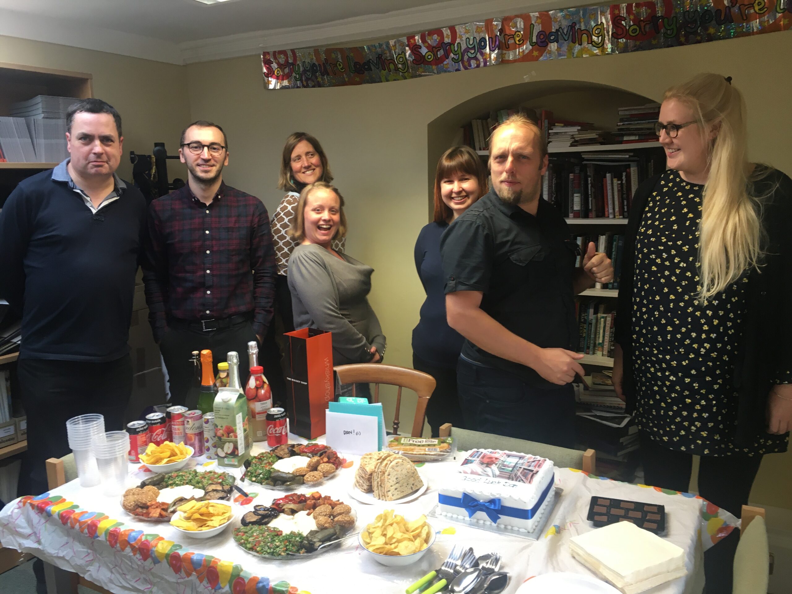 Full image of Dan poses with Bodleian coworkers in front of a party feast in a low-ceilinged office.