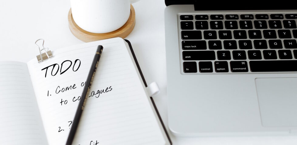Laptop and mug alongside a notebook and pencil. In the notebook is written "TODO: 1. Come out to colleagues. 2. ???. 3. Profit."