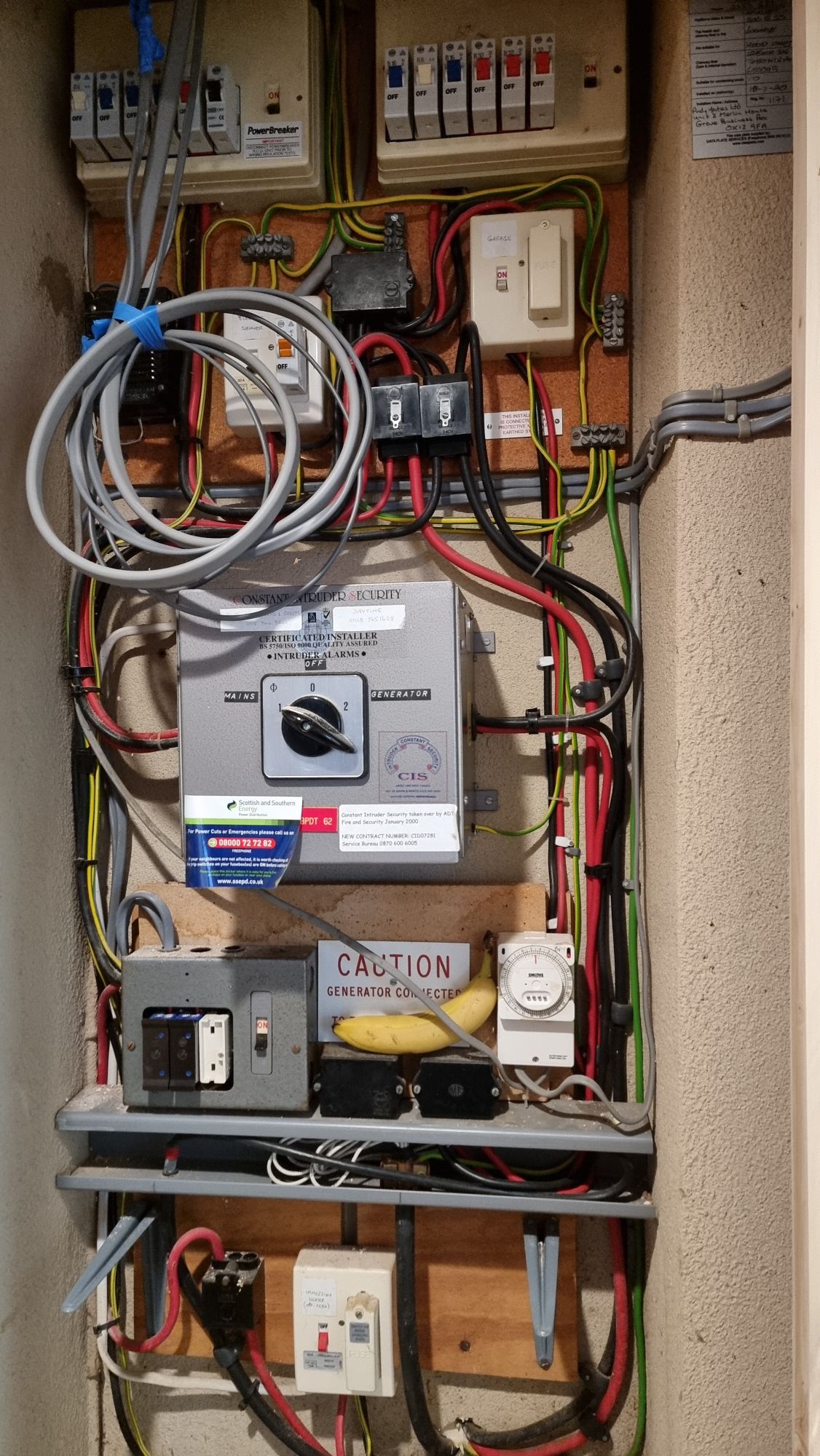 Full image of A cupboard the height of a fully-grown adult containing several fuseboxes (two large, one medium, and a handful of single-fuse ones, with a mixture of traditional fuses and RCD breakers), a large switch toggling between mains and a generator, a mains timer, and copious wiring. There's a banana in there too.