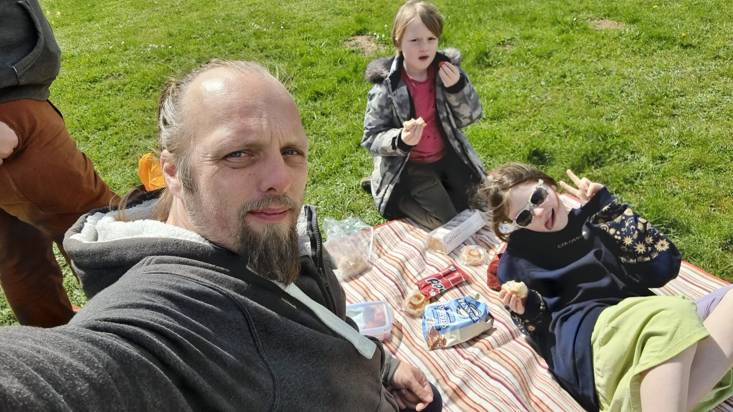 Full image of Dan kneels on a striped picnic mat with a 7-year-old and a 10-year old child alongside some sandwiches, iced fingers, Pepperami, fruit, and pretzels.