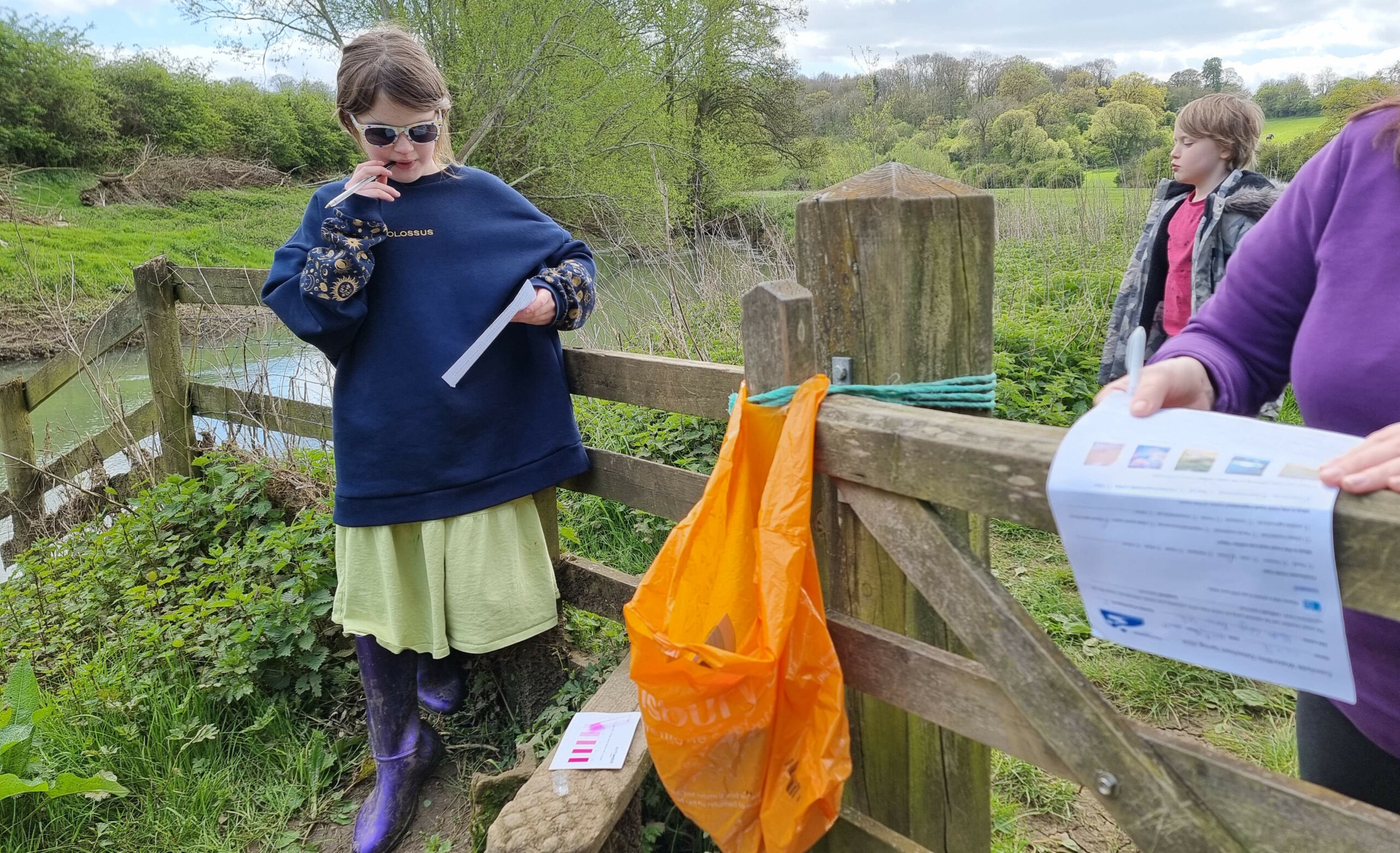 Full image of Children stand around at a riverside stile while a colour-changing chemical in a vial does its thing.