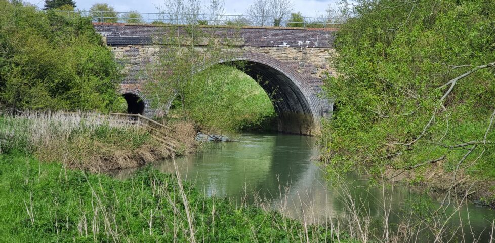 A brick viaduct with a single main span and two smaller spans crosses the River Evenlode, flanked with verdant grassy banks and trees with Spring buds and leaves.