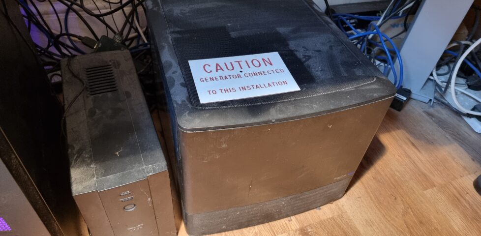 A cube-shaped black computer sits next to a battery pack on a laminated floor. A sign has been left atop it, reading "Caution: Generator connected to this installation."