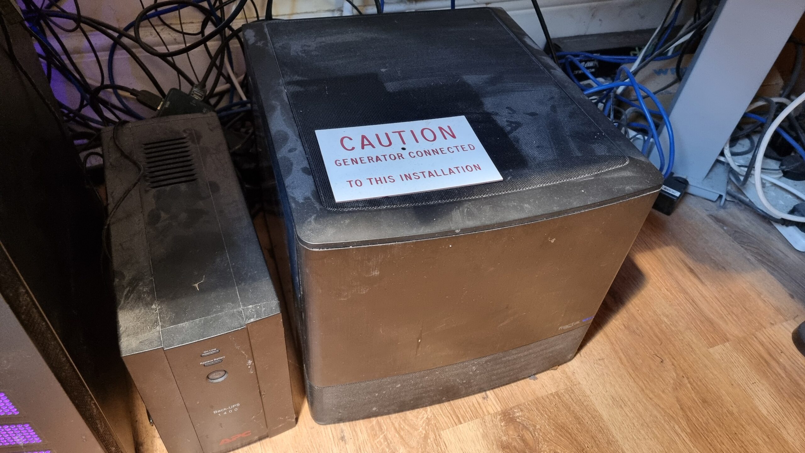 Full image of A cube-shaped black computer sits next to a battery pack on a laminated floor. A sign has been left atop it, reading "Caution: Generator connected to this installation."
