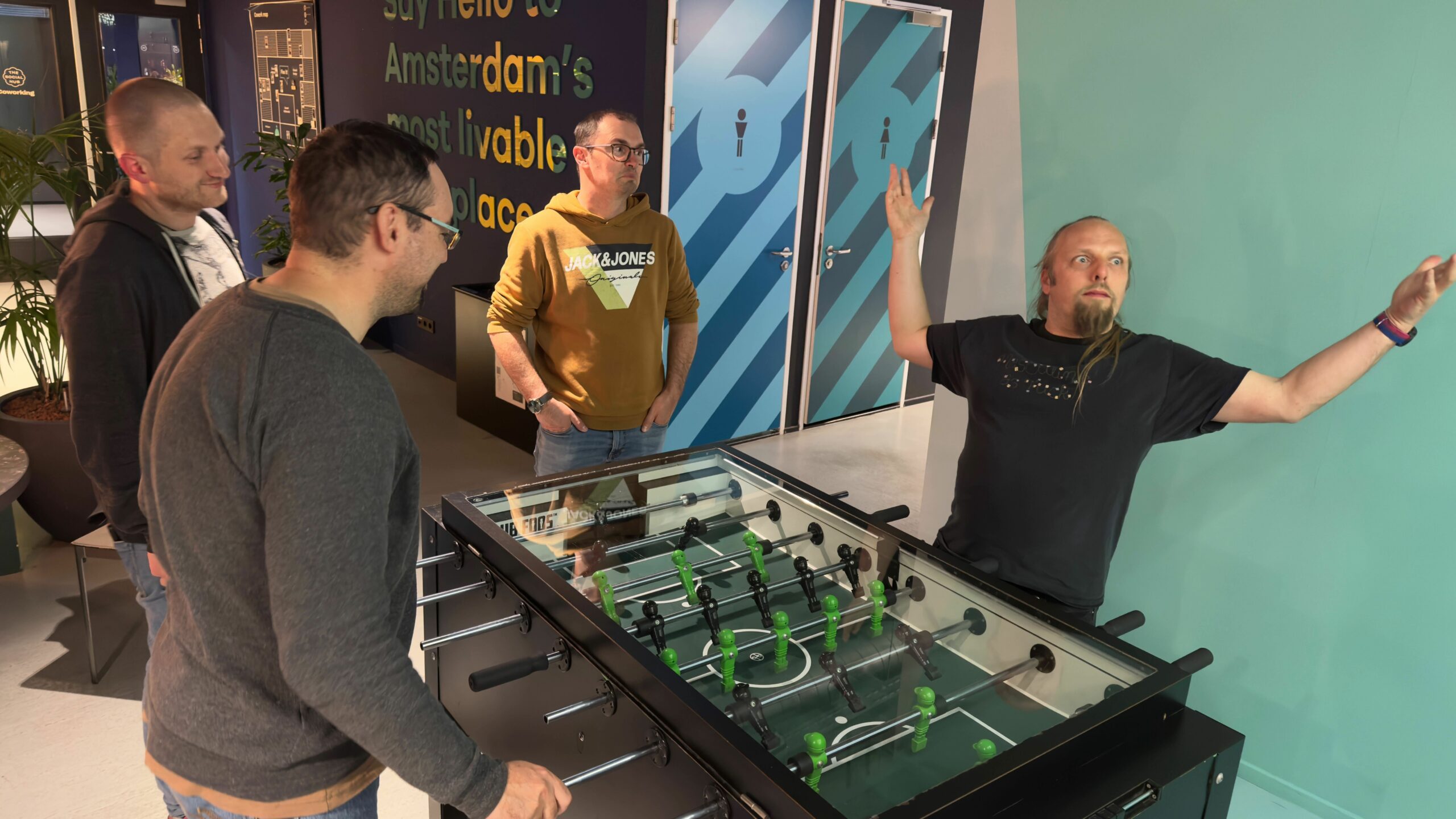 Full image of Dan, by a game of table football, throws his arms into the air as if in self-celebration.
