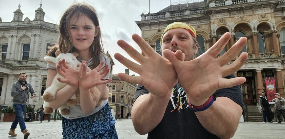Dan, wearing a black t-shirt and a rainbow-striped bandana, stands alongside a 10-year old girl carrying stuffed toys, in a market square. Both are making "spider" figures with their fingers, towards the camera.