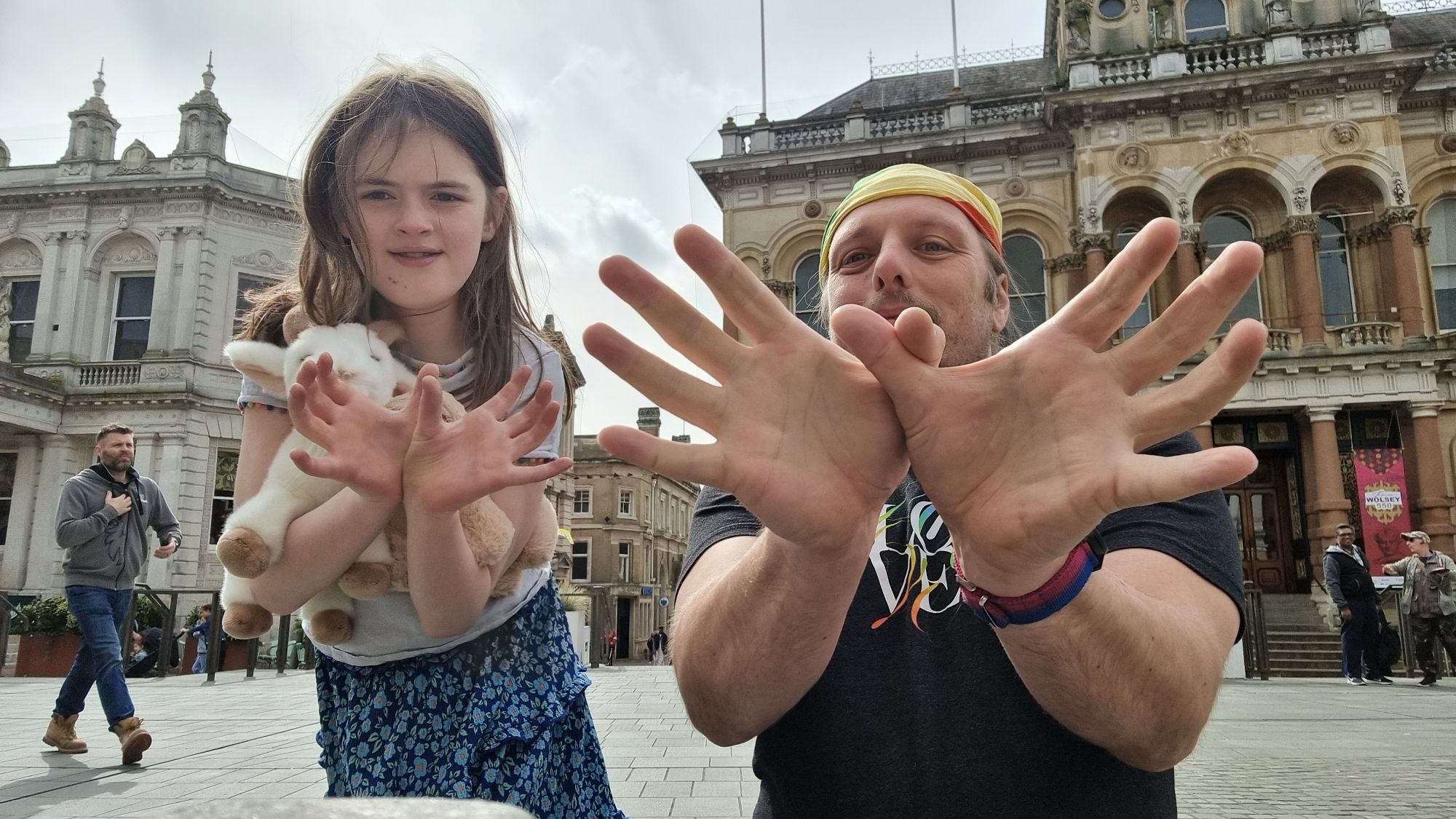 Full image of Dan, wearing a black t-shirt and a rainbow-striped bandana, stands alongside a 10-year old girl carrying stuffed toys, in a market square. Both are making "spider" figures with their fingers, towards the camera.